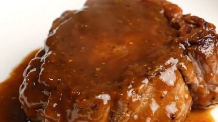 A close-up of a bone-in pork shoulder steak coated in a thick, glossy BBQ sauce inside a black slow cooker.