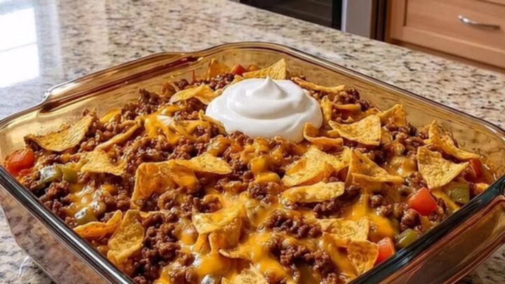 A warm, cheesy walking taco bake in a glass dish topped with sour cream and crushed chips on a granite counter.