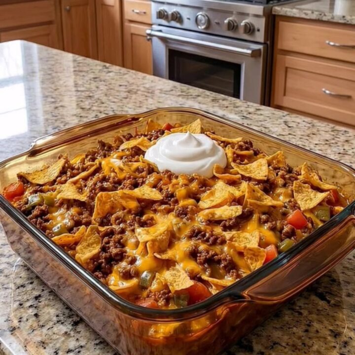 A warm, cheesy walking taco bake in a glass dish topped with sour cream and crushed chips on a granite counter.