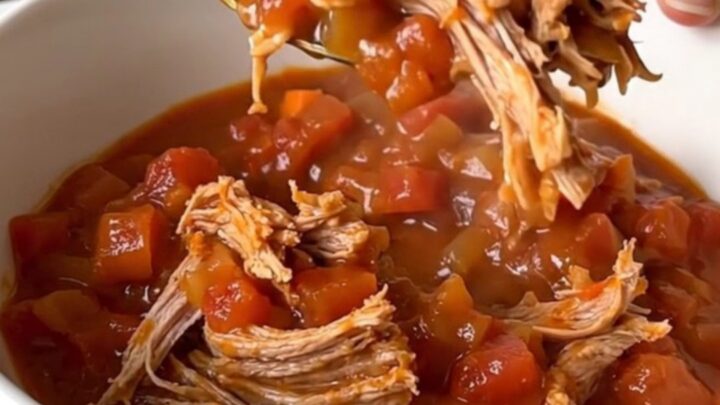 A close-up of a slow cooker filled with chunks of pork shoulder, diced carrots, onions, and tomato sauce.