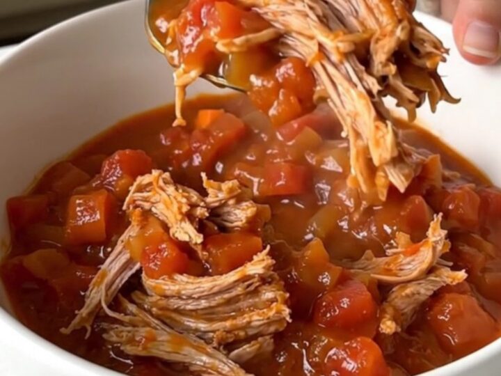 A close-up of a slow cooker filled with chunks of pork shoulder, diced carrots, onions, and tomato sauce.