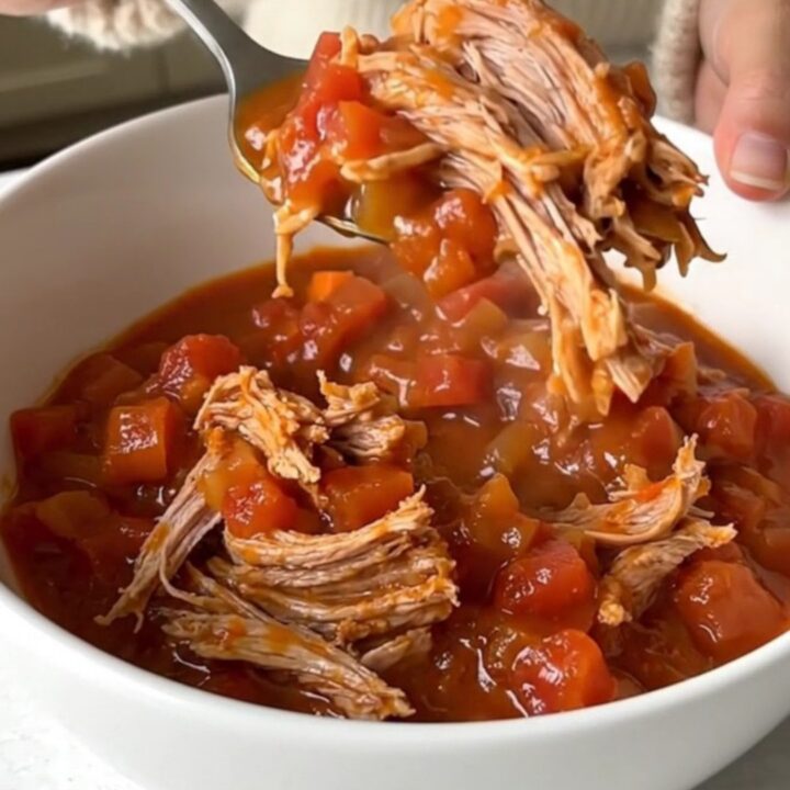 A close-up of a slow cooker filled with chunks of pork shoulder, diced carrots, onions, and tomato sauce.