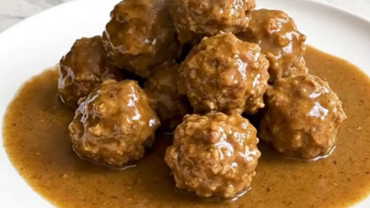 A serving spoon lifting juicy beef meatballs covered in creamy brown gravy from a black slow cooker.