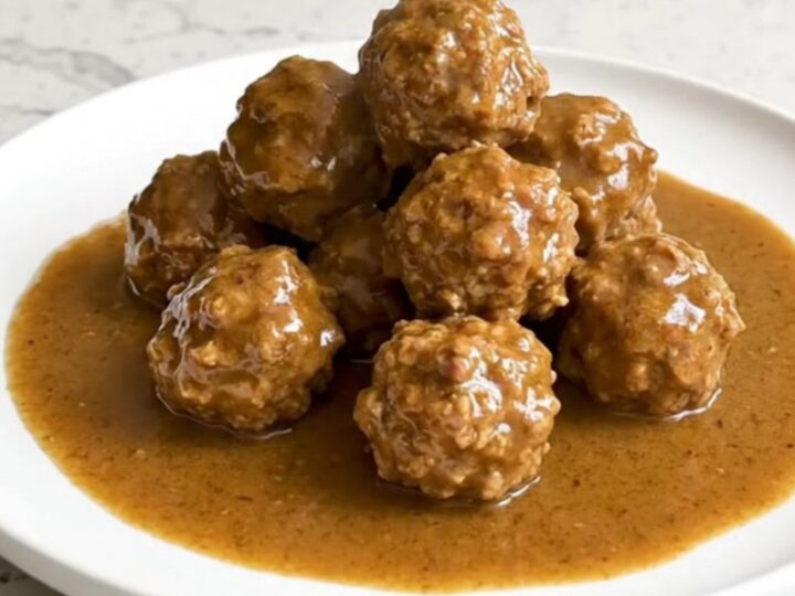 A serving spoon lifting juicy beef meatballs covered in creamy brown gravy from a black slow cooker.