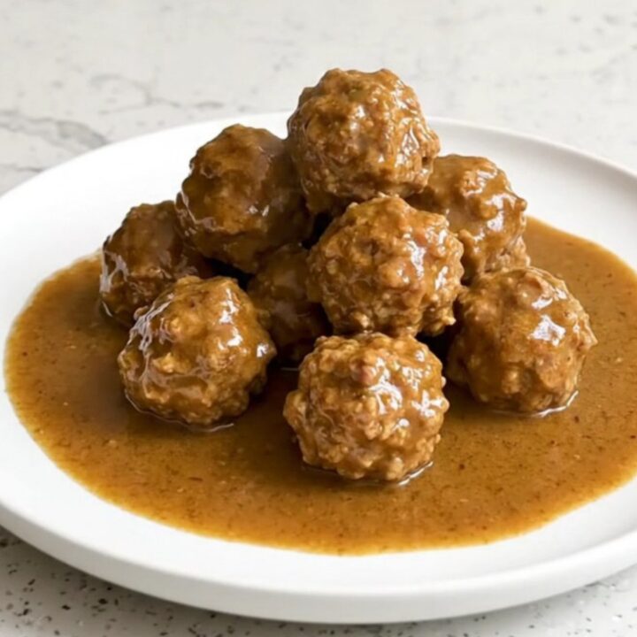 A serving spoon lifting juicy beef meatballs covered in creamy brown gravy from a black slow cooker.