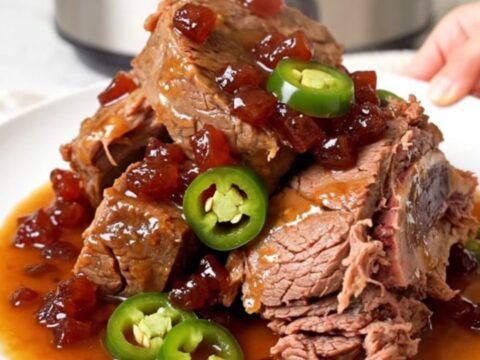 A whole, tender beef chuck roast sitting in a black slow cooker with a glossy pepper jelly and jalapeno sauce.