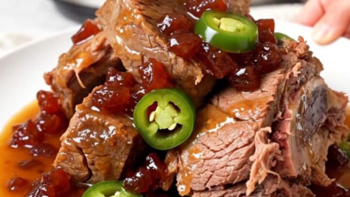A whole, tender beef chuck roast sitting in a black slow cooker with a glossy pepper jelly and jalapeno sauce.