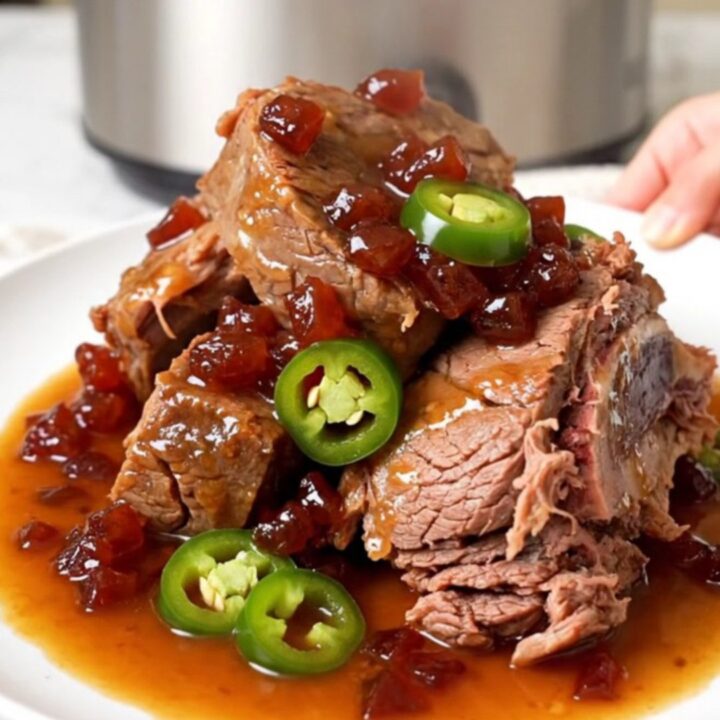 A whole, tender beef chuck roast sitting in a black slow cooker with a glossy pepper jelly and jalapeno sauce.