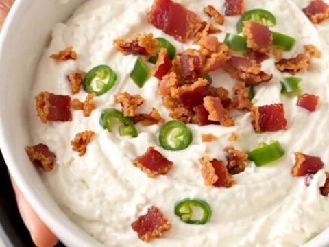 A close-up of creamy white queso dip in a black slow cooker topped with crispy bacon and diced green chiles.