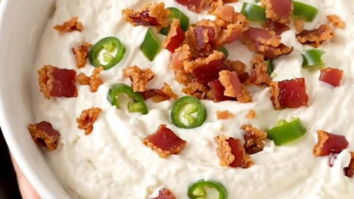 A close-up of creamy white queso dip in a black slow cooker topped with crispy bacon and diced green chiles.