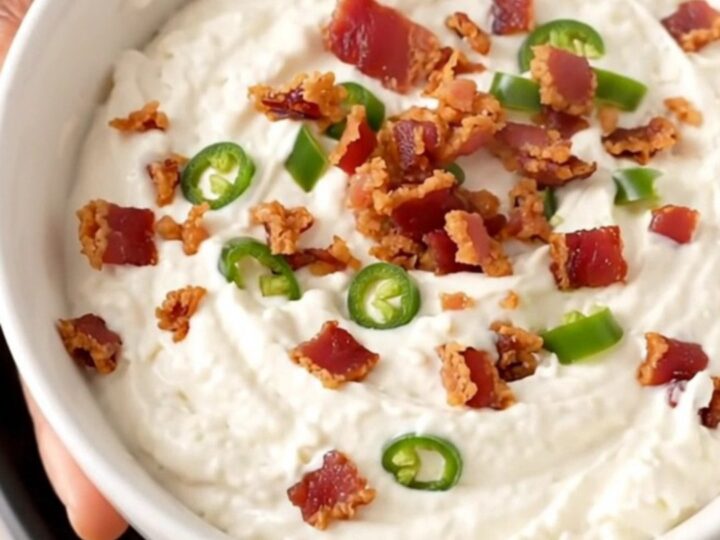 A close-up of creamy white queso dip in a black slow cooker topped with crispy bacon and diced green chiles.