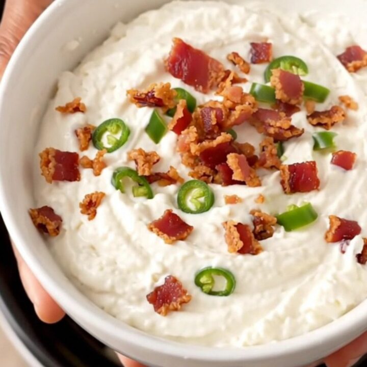 A close-up of creamy white queso dip in a black slow cooker topped with crispy bacon and diced green chiles.