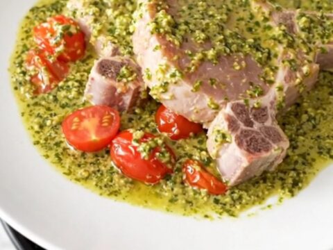 A close-up of creamy pesto pork chops served on a white plate with soft cherry tomatoes and a rich, green pesto sauce.