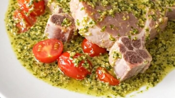A close-up of creamy pesto pork chops served on a white plate with soft cherry tomatoes and a rich, green pesto sauce.