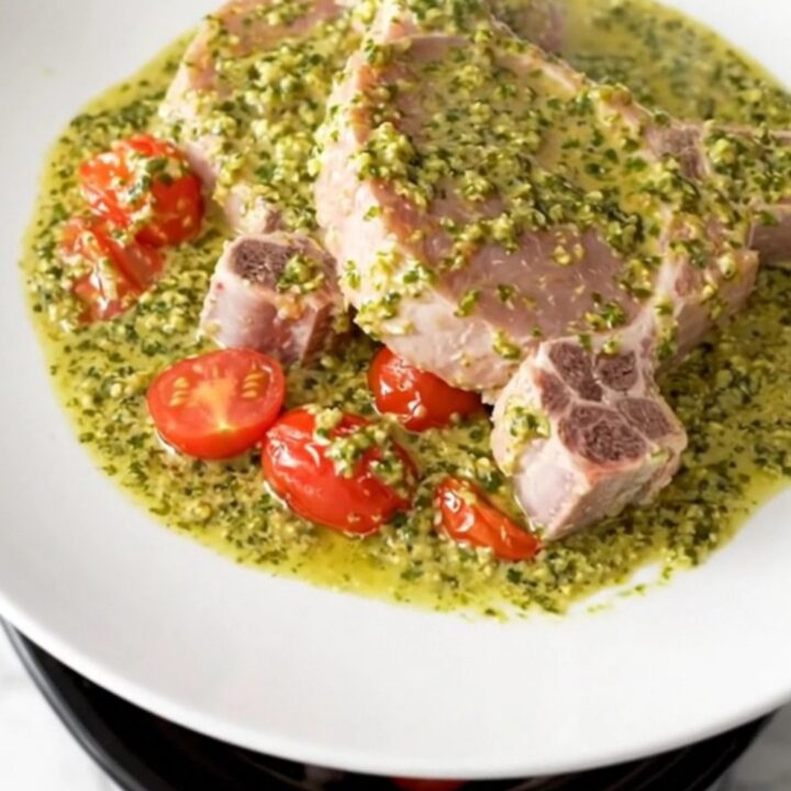 A close-up of creamy pesto pork chops served on a white plate with soft cherry tomatoes and a rich, green pesto sauce.