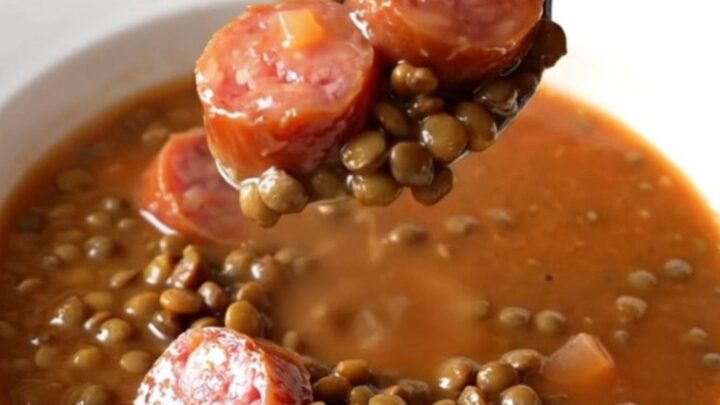 A close-up of a slow cooker filled with a hearty, colorful lentil and smoked sausage soup.