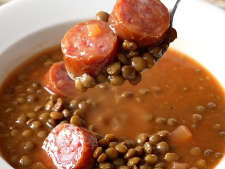 A close-up of a slow cooker filled with a hearty, colorful lentil and smoked sausage soup.