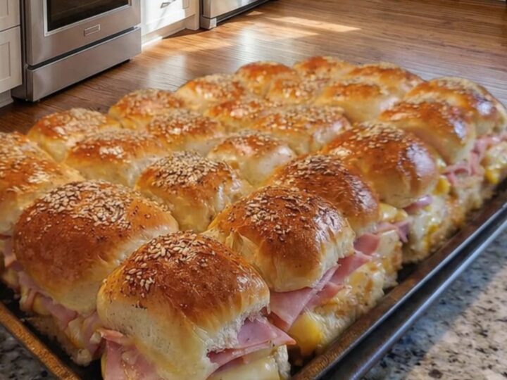 A tray of golden-brown baked ham and cheese sliders on a granite counter, with visible steam and melted cheese.