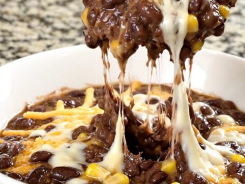 A black slow cooker filled with a creamy black bean, corn, and melted cheese dip on a granite countertop.