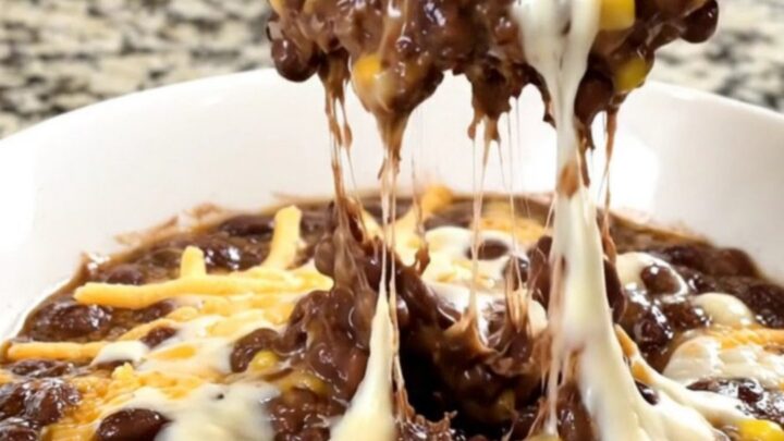 A black slow cooker filled with a creamy black bean, corn, and melted cheese dip on a granite countertop.