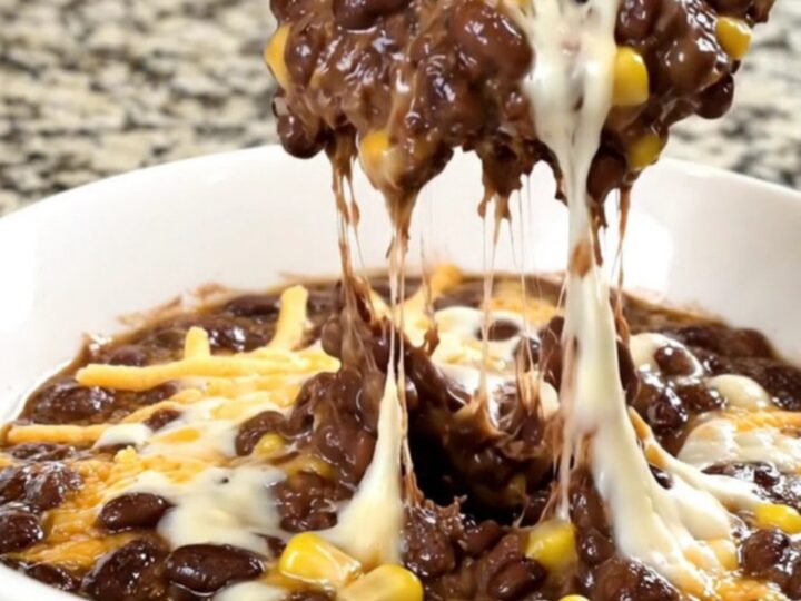 A black slow cooker filled with a creamy black bean, corn, and melted cheese dip on a granite countertop.
