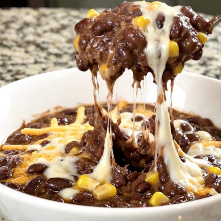 A black slow cooker filled with a creamy black bean, corn, and melted cheese dip on a granite countertop.