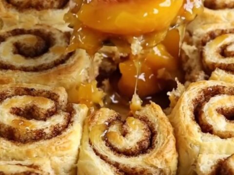 A warm cinnamon roll peach cobbler in a white ceramic dish with golden dough pieces.