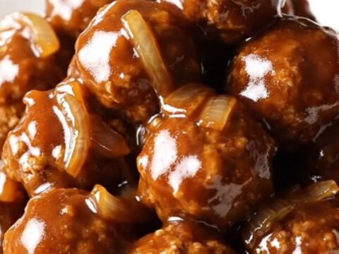 A close-up of tender meatloaf meatballs coated in a glossy, sweet, and tangy ketchup glaze with soft onions in a slow cooker.