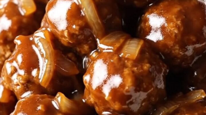 A close-up of tender meatloaf meatballs coated in a glossy, sweet, and tangy ketchup glaze with soft onions in a slow cooker.