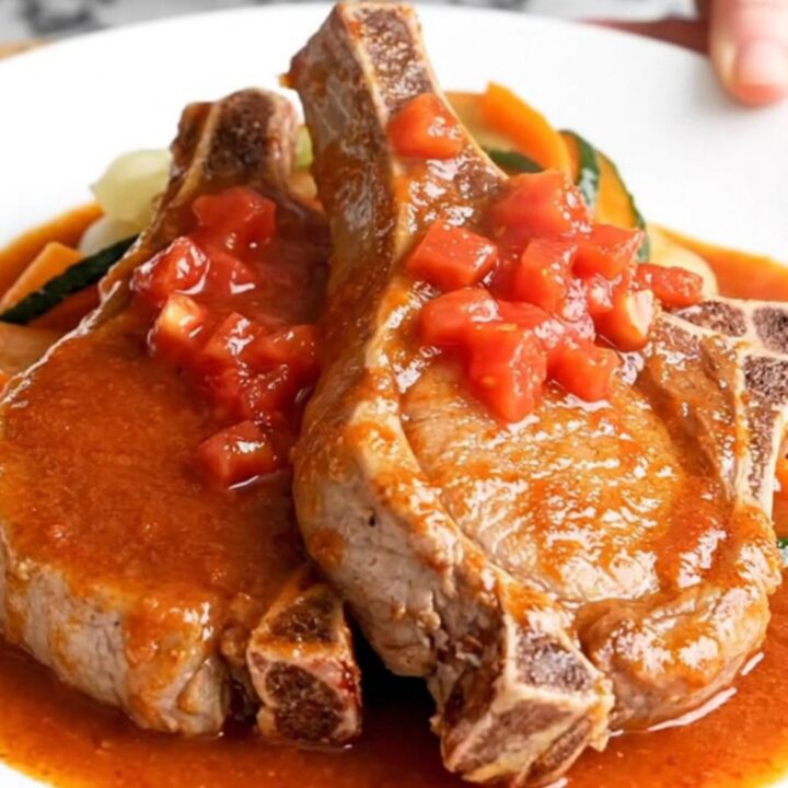 A top-down view of raw bone-in pork chops in a white slow cooker, topped with diced vegetables and Creole seasoning.