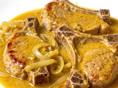 A close-up of tender, braised pork chops covered in a creamy, golden beer and cheddar cheese sauce on a white plate.