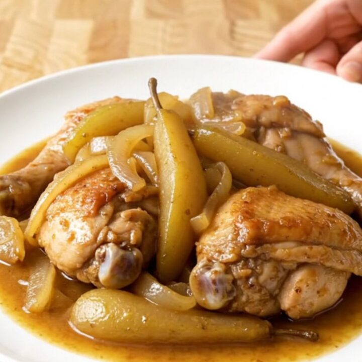 A close-up of tender, slow-cooked chicken thighs topped with soft, spiced pears and onions in a stainless steel slow cooker.