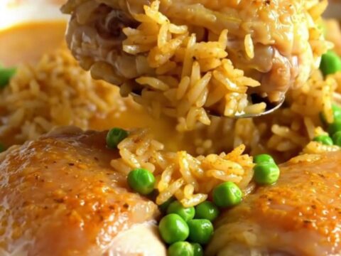 A top-down view of a rectangular baking dish filled with seasoned rice, green peas, and tender chicken thighs.