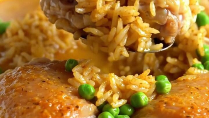 A top-down view of a rectangular baking dish filled with seasoned rice, green peas, and tender chicken thighs.