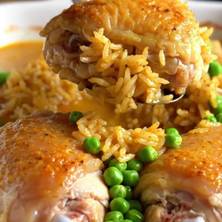 A top-down view of a rectangular baking dish filled with seasoned rice, green peas, and tender chicken thighs.