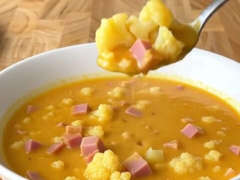 A close-up shot of a white bowl filled with creamy cheesy ham and cauliflower soup, with a spoon lifting a bite.