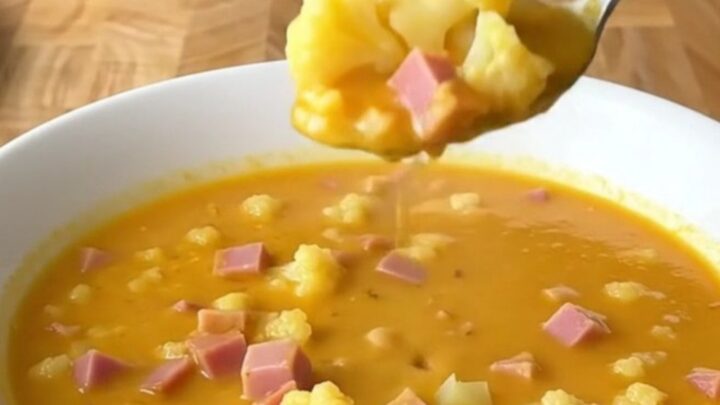 A close-up shot of a white bowl filled with creamy cheesy ham and cauliflower soup, with a spoon lifting a bite.