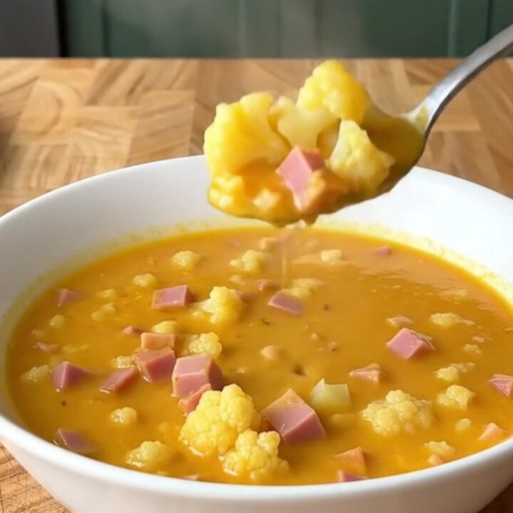 A close-up shot of a white bowl filled with creamy cheesy ham and cauliflower soup, with a spoon lifting a bite.