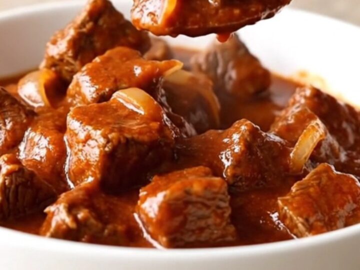 A close-up of tender beef cubes in a rich red chile sauce inside a white bowl.
