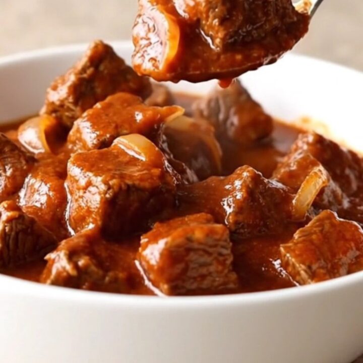 A close-up of tender beef cubes in a rich red chile sauce inside a white bowl.