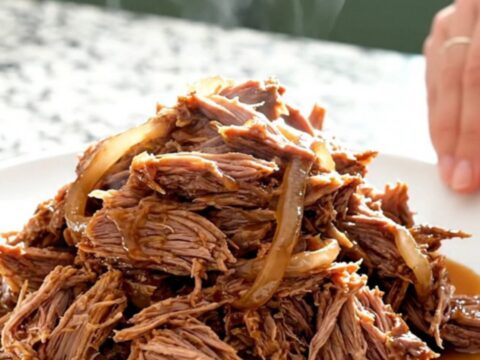 A close-up of tender, shredded pulled pork mounded on a white plate with visible smoky juices and soft onions.