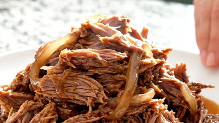 A close-up of tender, shredded pulled pork mounded on a white plate with visible smoky juices and soft onions.
