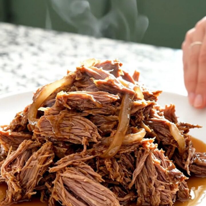 A close-up of tender, shredded pulled pork mounded on a white plate with visible smoky juices and soft onions.