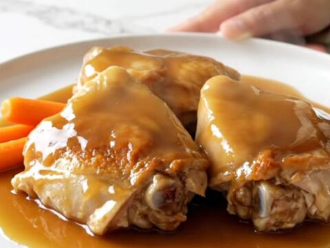 A top-down view of six raw bone-in chicken thighs and baby carrots in a slow cooker, being drizzled with maple syrup.