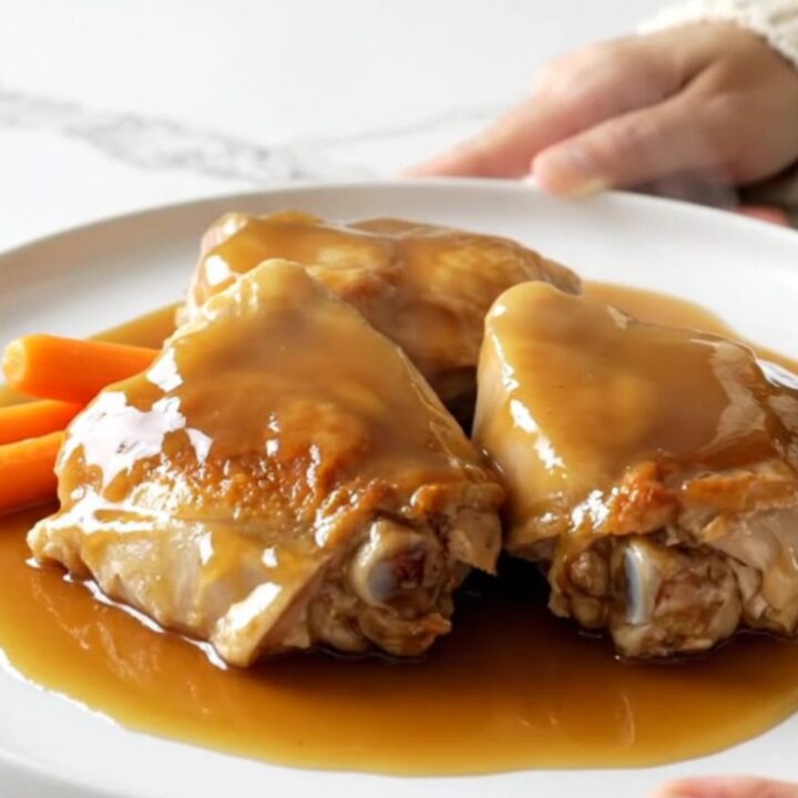 A top-down view of six raw bone-in chicken thighs and baby carrots in a slow cooker, being drizzled with maple syrup.