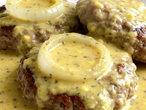 A top-down view of raw turkey burger steaks arranged in a slow cooker with onions and creamy Dijon sauce.