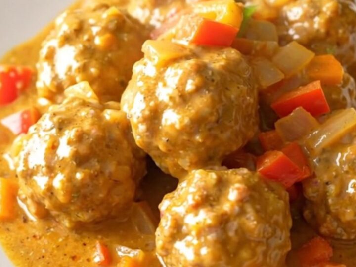 A close-up of creamy Cajun meatballs in a slow cooker topped with colorful bell peppers and onions.