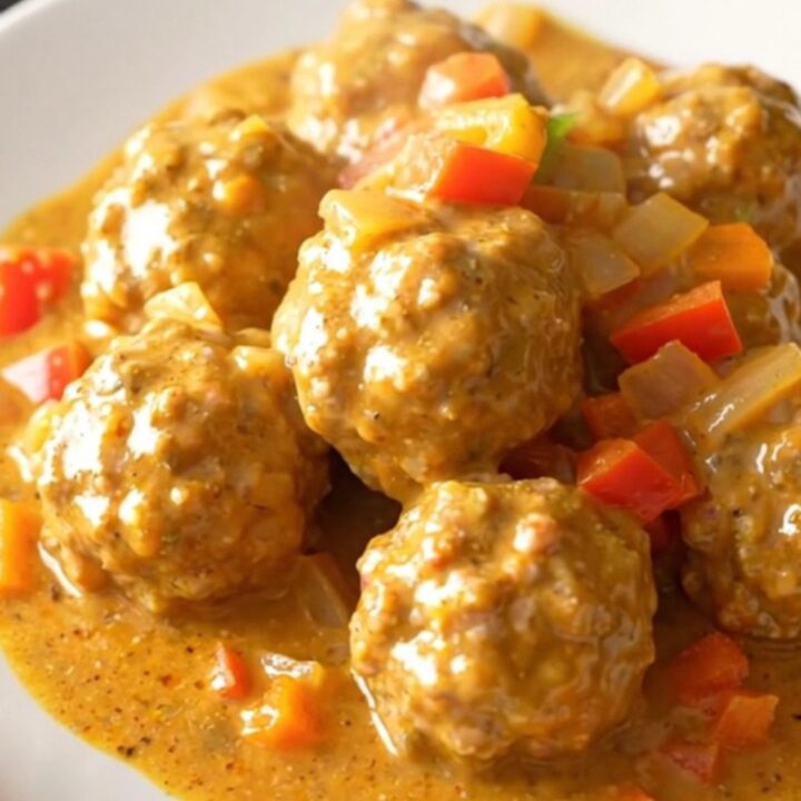A close-up of creamy Cajun meatballs in a slow cooker topped with colorful bell peppers and onions.