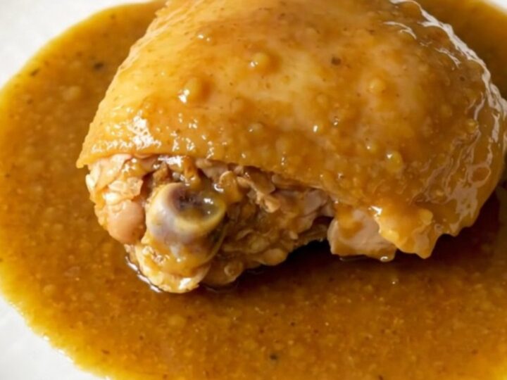 A close-up view of slow-cooked chicken thighs coated in a thick honey curry sauce with soft onions on a white plate.