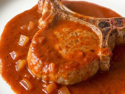 Tender pork chops smothered in vibrant red enchilada sauce with soft onions, served on a white plate.